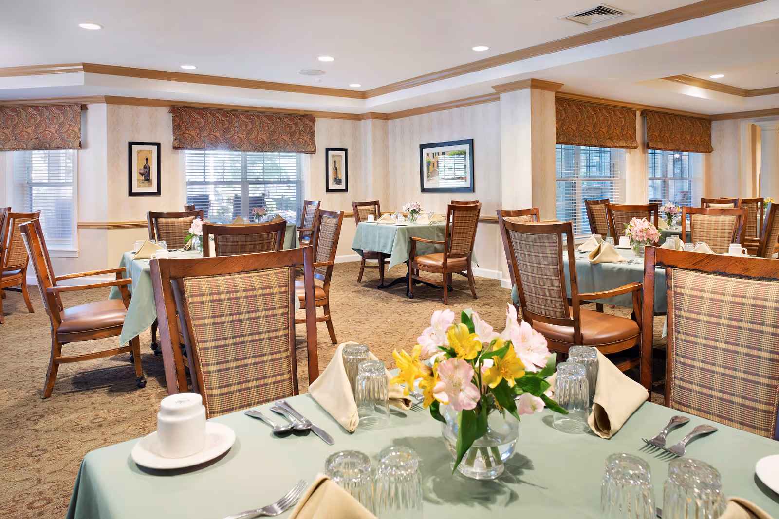 A well-lit dining room with multiple tables covered in light green tablecloths, each set with beige napkins, glassware, silverware, and floral centerpieces. The room features wooden chairs with plaid upholstery, large windows with patterned valances, framed artwork on the walls, and a carpeted floor.