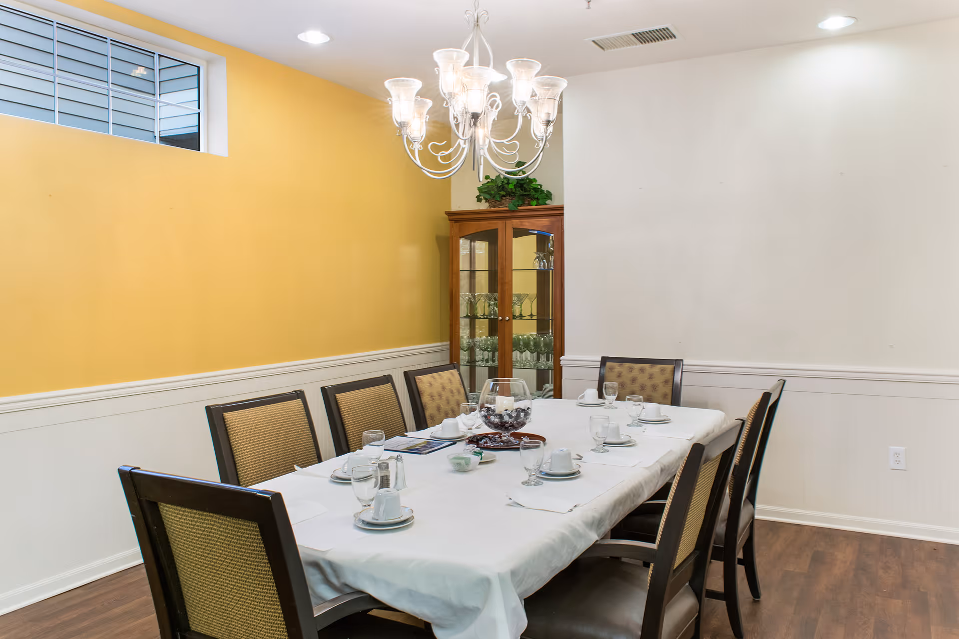 A dining room with a rectangular table covered with a white tablecloth set for eight people. The table has cups, saucers, glasses, and a centerpiece. There are eight chairs around the table. The walls are painted yellow and white, and there is a glass-fronted wooden cabinet with glassware in the corner. A chandelier with multiple lights hangs above the table.