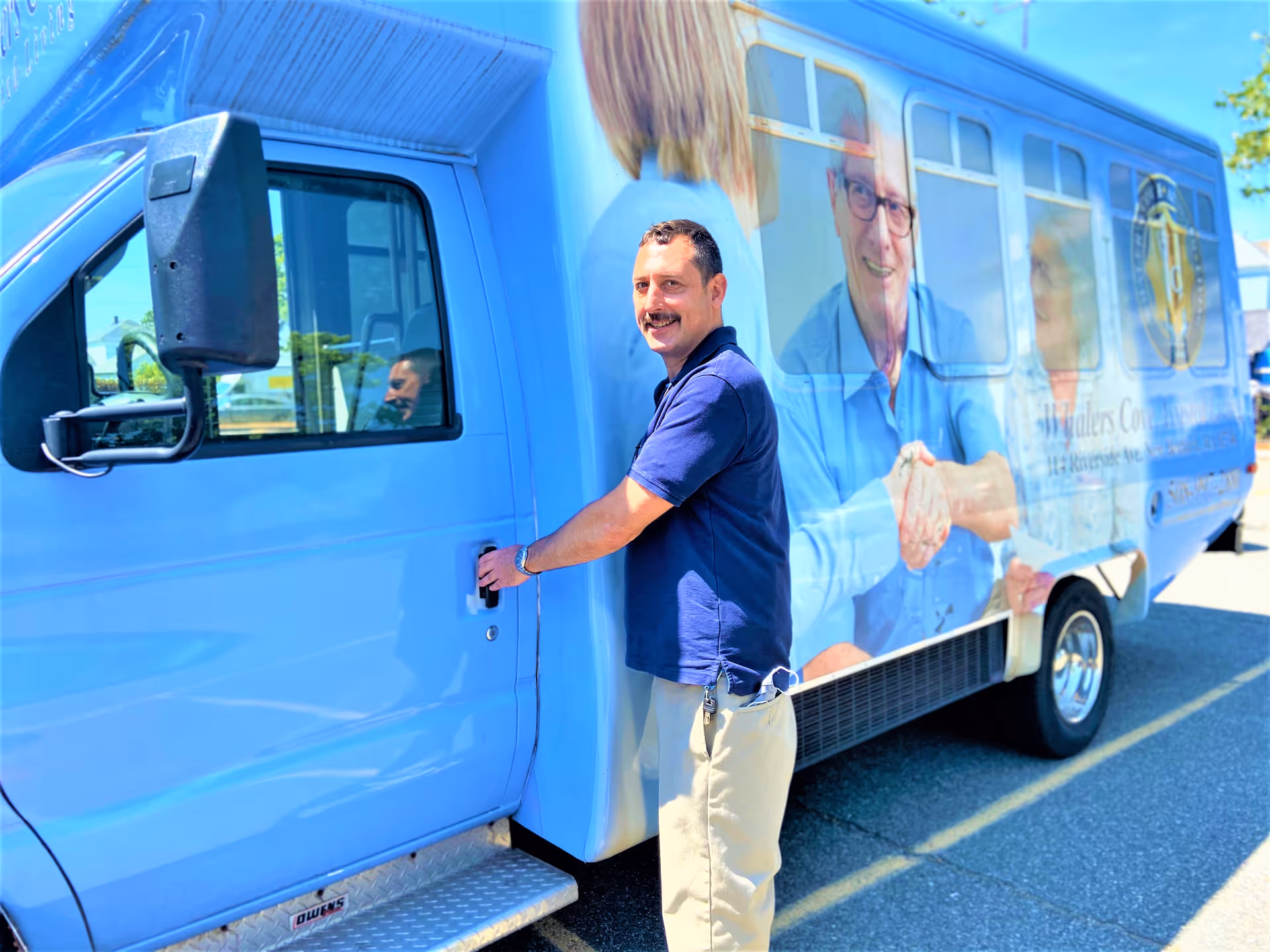 A man in a navy blue polo shirt and beige pants is standing outside a blue shuttle bus, holding the door handle and smiling. The bus has large images of elderly people and the logo of Whaler's Cove Assisted Living on its side.