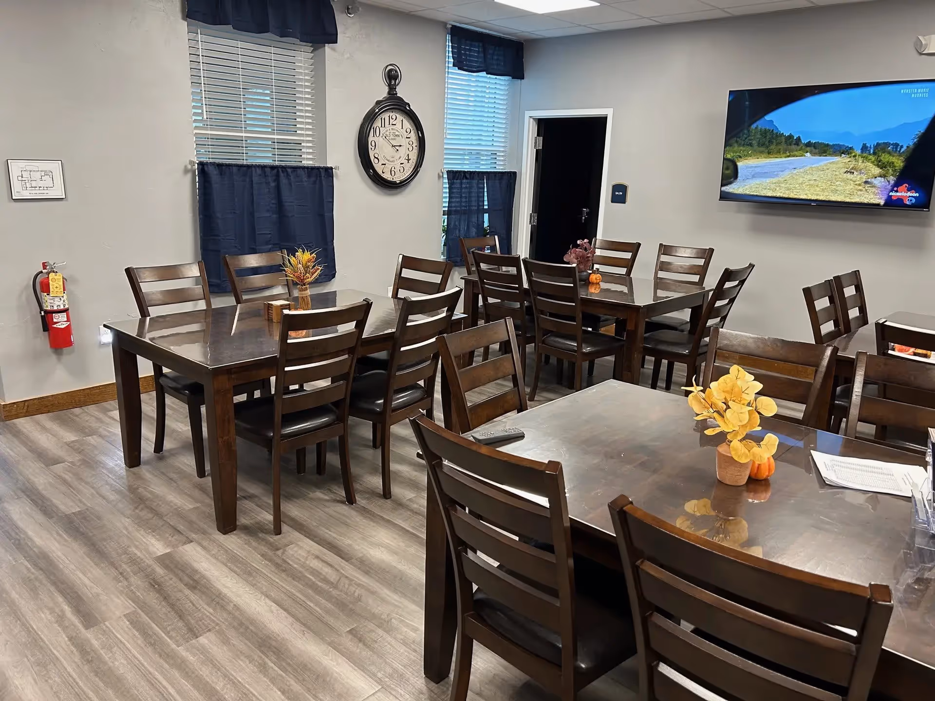 A dining room with three wooden tables and multiple wooden chairs arranged around them. Each table has a small decorative centerpiece with flowers and pumpkins. The room has light gray walls, two windows with blue curtains, a large wall clock, a fire extinguisher mounted on the wall, and a flat-screen TV displaying a nature scene.