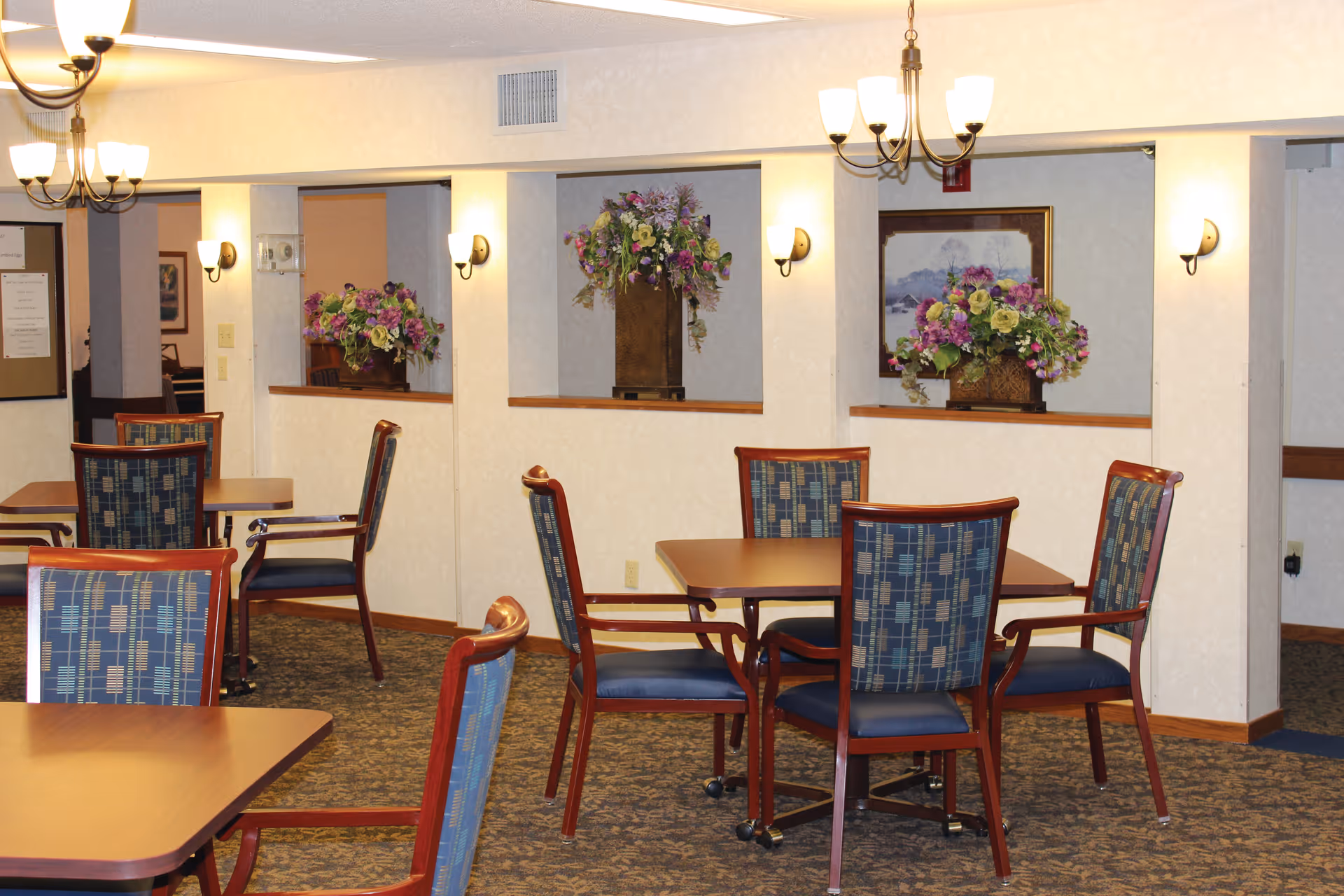 Dining room with several tables and upholstered chairs, floral arrangements on ledges, chandeliers and wall sconces.