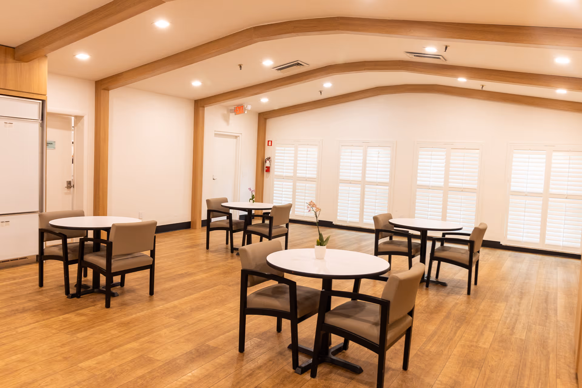 A bright dining room with wooden floors and white walls featuring four round tables, each surrounded by four cushioned chairs. Each table has a small potted plant as a centerpiece. The room has large windows with white shutters and wooden beams on the ceiling.