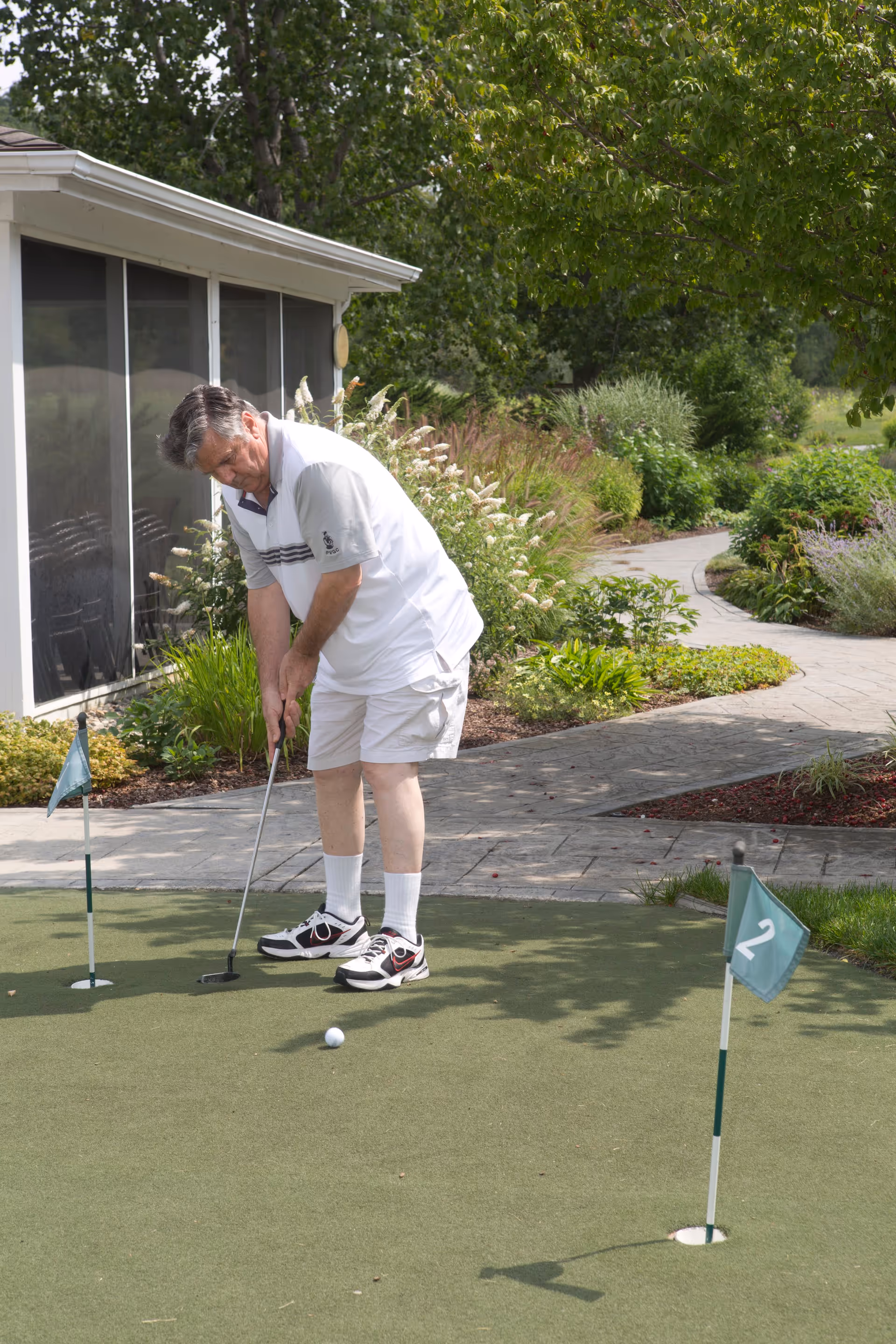 An elderly man wearing a white shirt, white shorts, and sneakers is playing putting golf on a small green putting area outdoors. The setting includes a paved walkway, lush greenery, and trees in the background.