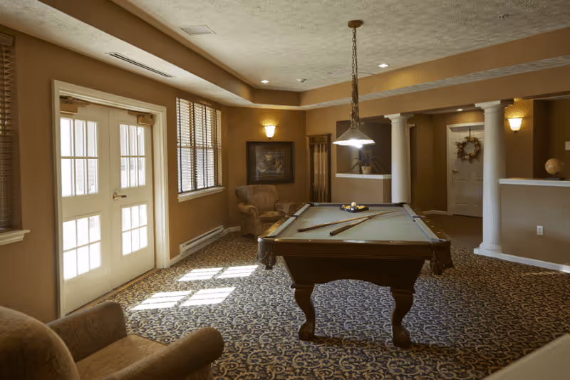 A cozy indoor recreation room featuring a pool table under a hanging light, upholstered chairs, columns, and sunlight streaming through double doors.