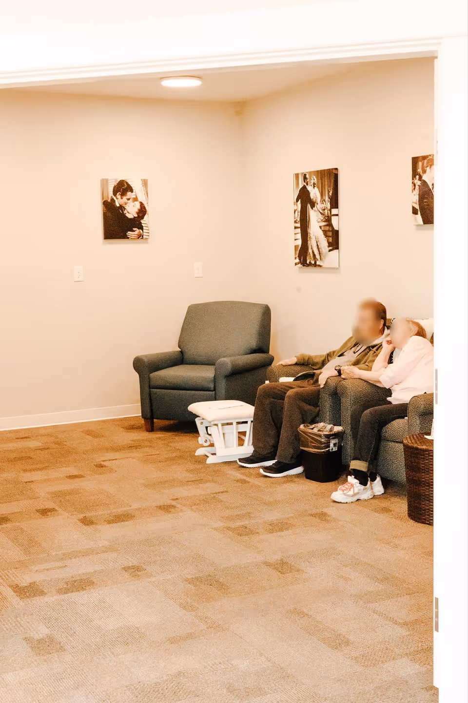 A cozy sitting area in a senior living facility with two elderly people sitting on a couch. There is an empty armchair and a small white footrest nearby. The walls are decorated with framed black and white photos of couples dancing and embracing. The carpet is patterned in neutral tones.