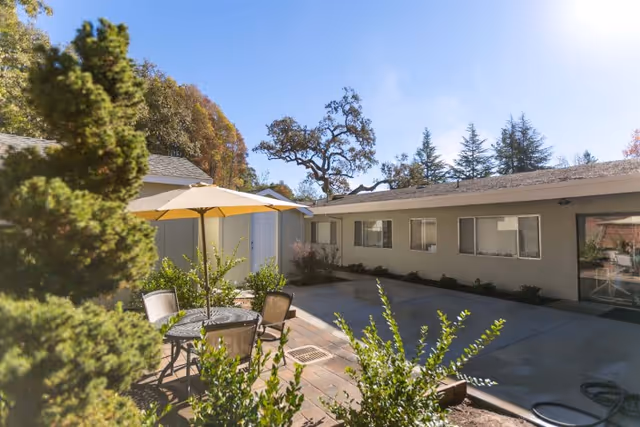 Sunny outdoor courtyard of a single-story care facility with a patio table, umbrella, chairs, and surrounding landscaping.