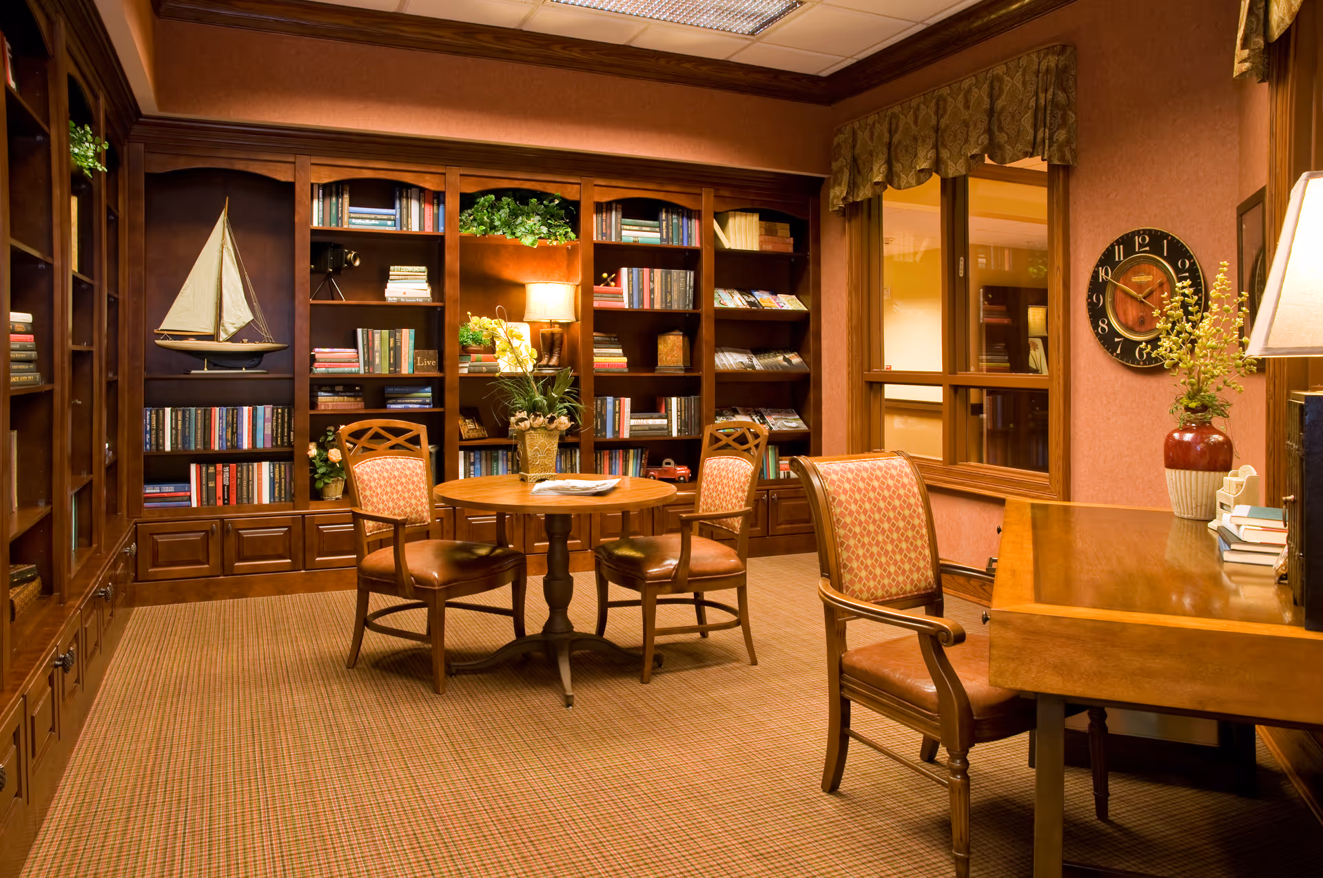 A cozy library room with wooden bookshelves filled with books and decorative items, a round wooden table with four cushioned chairs, a wooden desk with a lamp and a vase with flowers, and a wall clock. The room has warm lighting and a window with a valance.