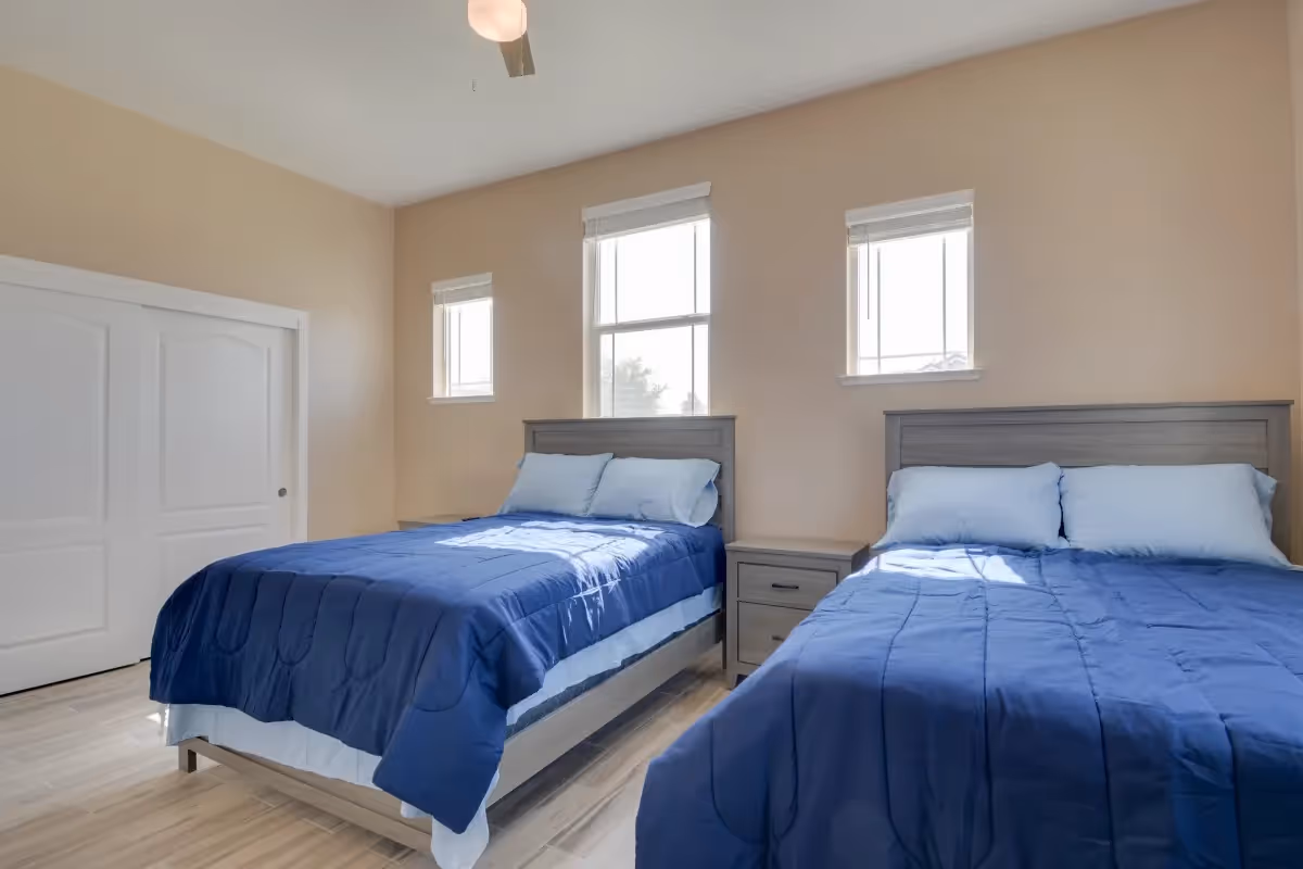 A bright bedroom with two twin beds featuring blue comforters and light blue pillows. Between the beds is a wooden nightstand with two drawers. The room has three windows with white blinds, beige walls, a ceiling fan with a light, and a white sliding closet door.