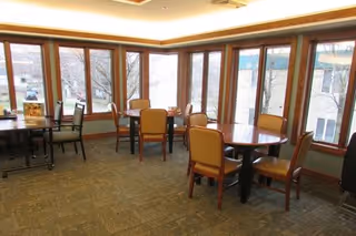 A bright dining area with multiple round and rectangular tables surrounded by chairs with tan upholstery. Large windows line the walls, allowing natural light to fill the room and providing a view of the outside buildings and trees.