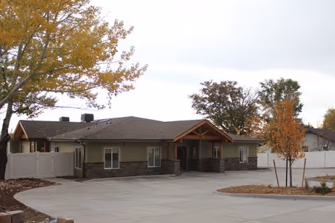 Single-story building with a pitched roof and a covered entrance, surrounded by a white fence and some trees with autumn foliage, under a cloudy sky.