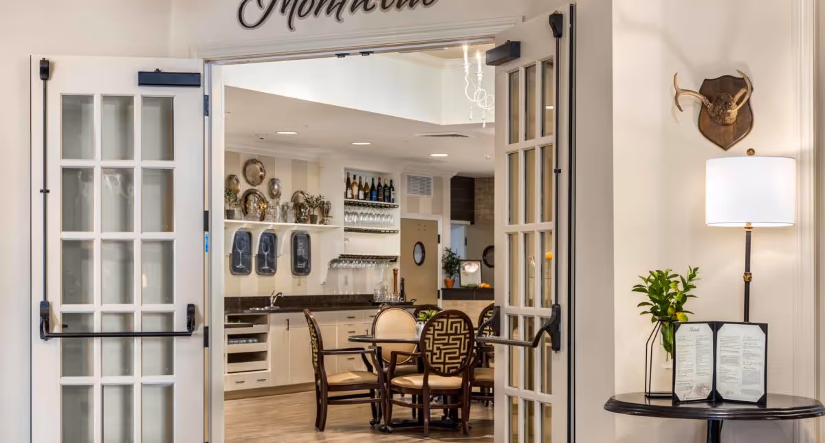 View through open double glass doors into a dining area with round tables and chairs. The dining area has shelves with decorative plates, wine bottles, and glasses. To the right, there is a small round table with a menu, a plant, a lamp, and a mounted deer head on the wall.