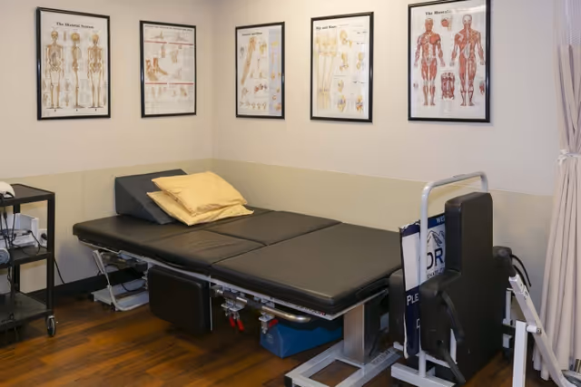 A rehabilitation room with a black padded treatment table, yellow pillows, anatomical charts of the skeletal and muscular systems on the walls, and medical equipment on a small cart and nearby stand.