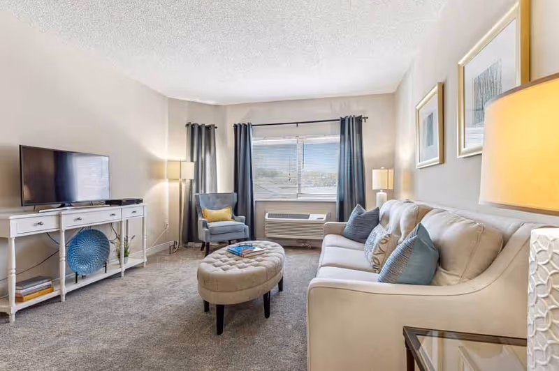 A cozy living room with a beige sofa adorned with blue and patterned pillows, a tufted oval ottoman in the center, a blue armchair with a yellow pillow near a window with blue curtains, a white TV stand with a flat-screen TV, and two table lamps providing warm lighting.