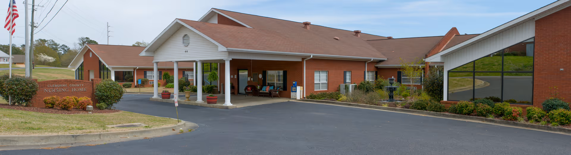 Front exterior view of Cleburne County Nursing Home building with a covered entrance, brick walls, large windows, and landscaped bushes. An American flag is visible on the left side near the facility sign.