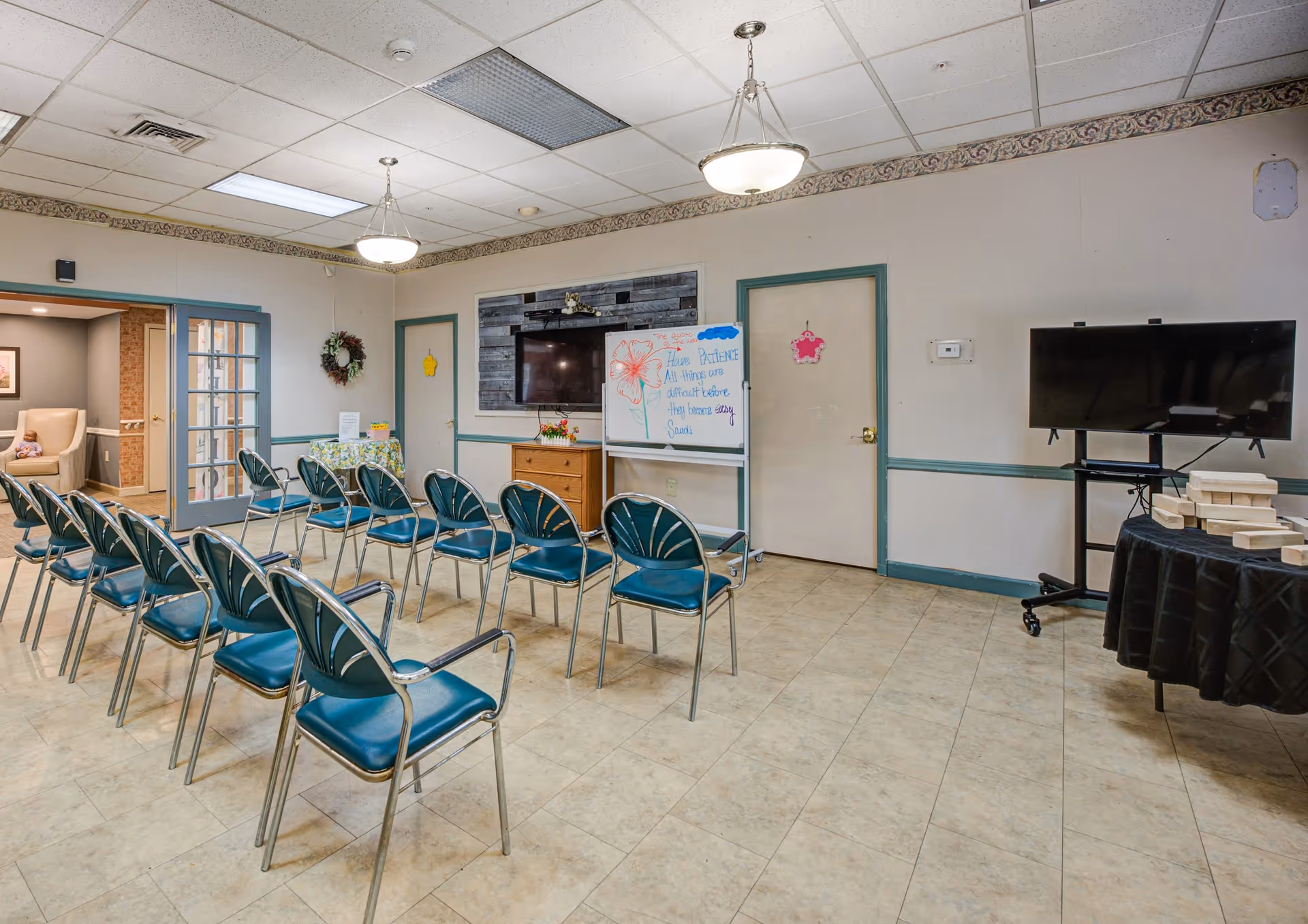 A community room with rows of green chairs arranged facing a wall-mounted TV and a whiteboard with a motivational message. The room has tiled floors, two hanging ceiling lights, and a table with wooden blocks on the right. There are two doors and a small table with a floral tablecloth and a wreath on the wall. A person is partially visible sitting in an adjacent room through a doorway on the left.