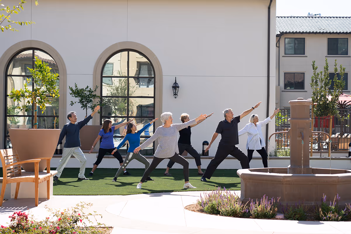 A group of seniors practicing yoga or stretching exercises outdoors on a grassy area in front of a building with large arched windows. There is a water fountain and some plants around the area, and the seniors are standing in a line with their arms extended.