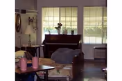 A cozy living room area with a piano against a wall of large windows. There is a floral-patterned armchair, a small table with pink candles, a lamp, and a bean bag chair in front of the piano. The room has soft natural light coming through the windows.