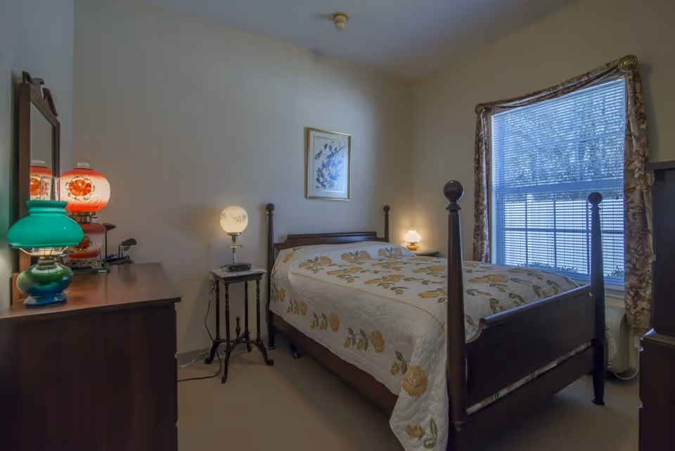 A cozy bedroom with a wooden four-poster bed covered with a floral quilt. There is a window with blinds and floral curtains on the right side. A wooden dresser with a mirror and several decorative lamps is on the left side of the room. A small side table with a lamp is next to the bed. The walls are light-colored and there is a framed picture hanging above the bed.