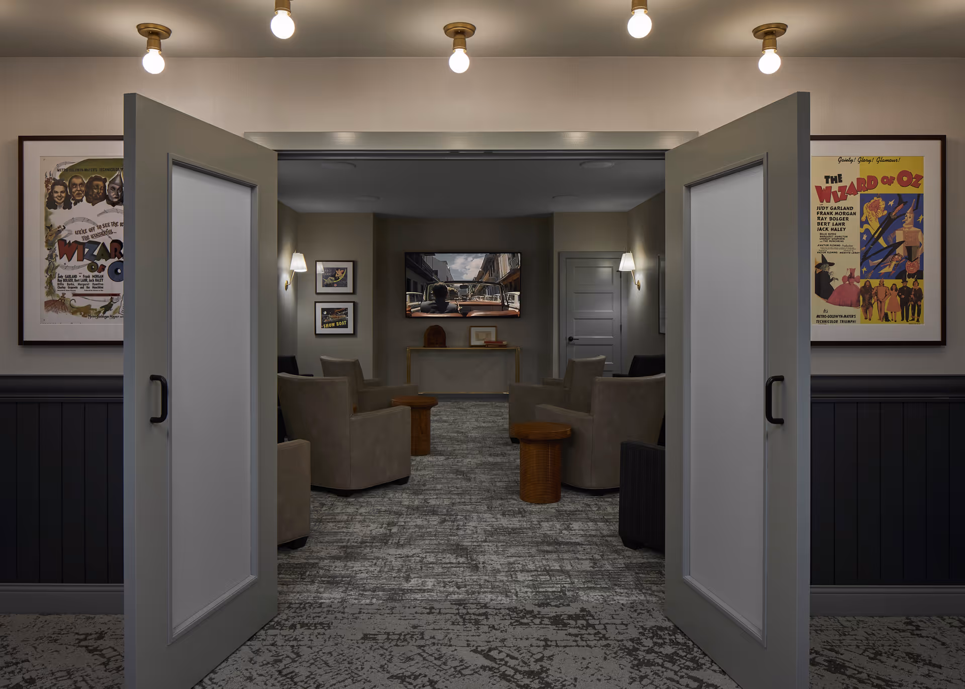 View through open double doors into a cozy living room with several beige armchairs arranged around small wooden tables. A flat-screen TV is mounted on the far wall displaying a street scene. The walls are decorated with framed posters, including two Wizard of Oz posters on either side of the doors. The room is softly lit by wall sconces and ceiling lights.