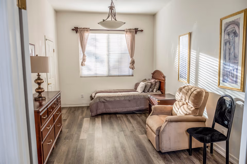 Small furnished bedroom with a single bed, dresser and lamp, recliner and window with blinds.