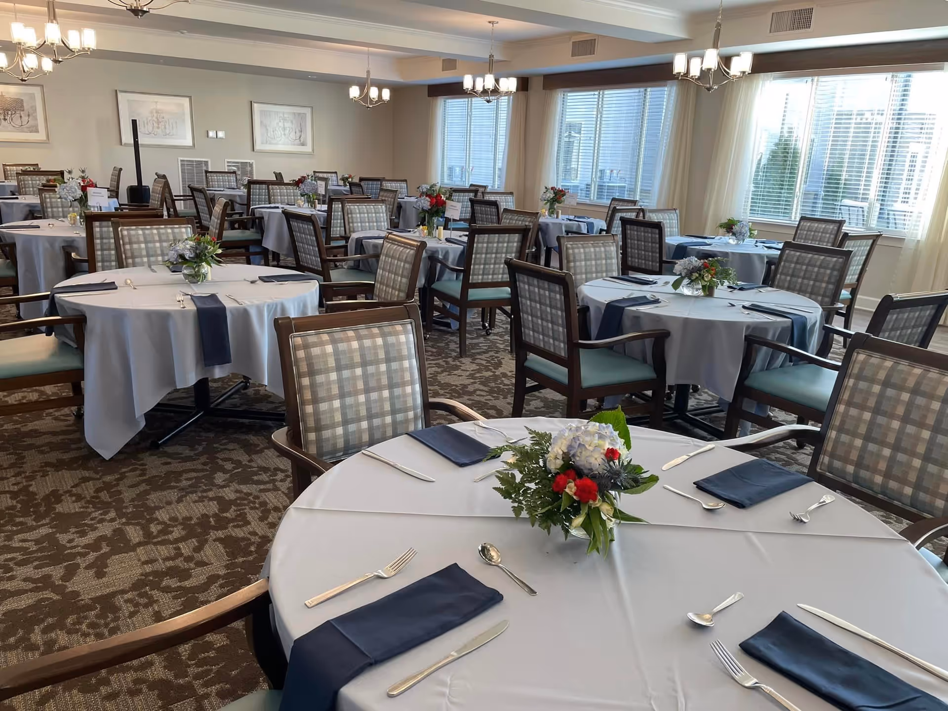 Bright dining room with multiple round tables set with white tablecloths, navy napkins, floral centerpieces, and plaid-upholstered chairs.