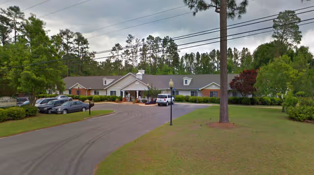 Single-story senior living facility building with a circular driveway, parked cars, and surrounding trees and lawn.