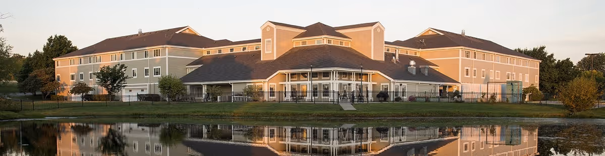Wide exterior view of Fort Scott Presbyterian Village, a large multi-story senior living facility with beige and light green walls, multiple windows, and a central entrance. The building is reflected in a calm pond in the foreground, with trees and greenery surrounding the area under a clear sky.