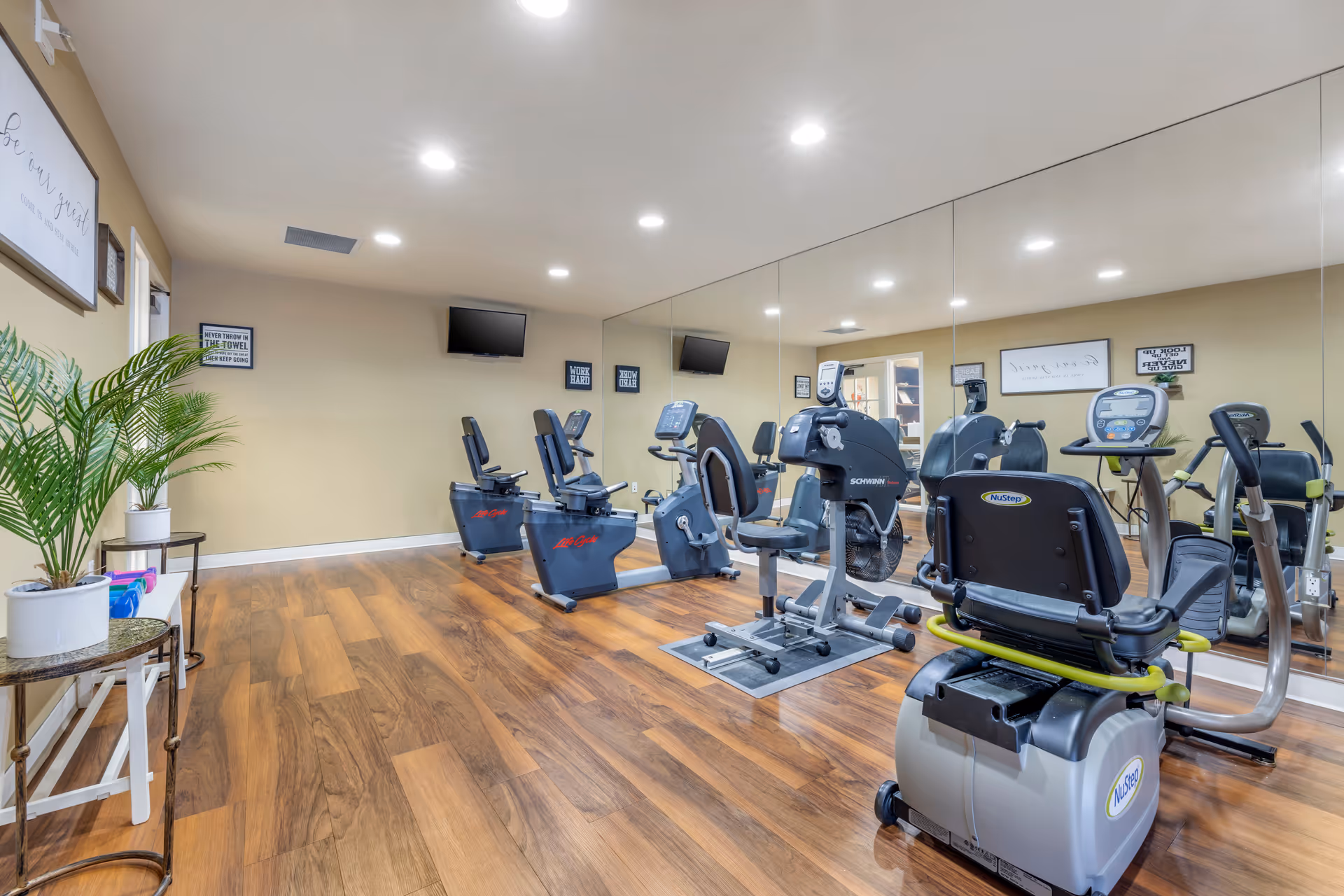 A fitness room with several exercise bikes and cardio machines on a wooden floor. The walls are beige with motivational signs and a flat-screen TV mounted on one wall. One wall is covered with large mirrors reflecting the equipment. There are also some potted plants and small tables along one side of the room.