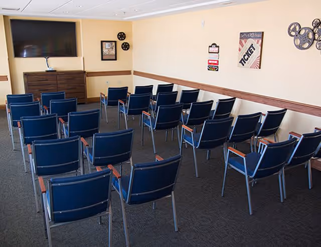 Room with multiple rows of blue chairs facing a large flat screen TV mounted on a beige wall. The room has movie-themed decorations including film reels and a ticket sign on the walls.