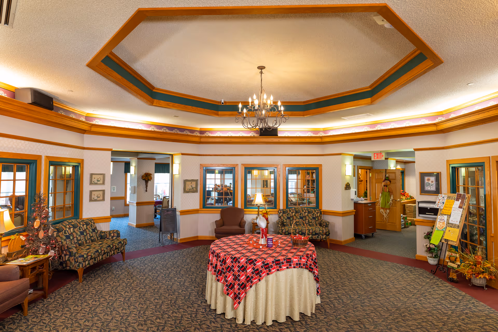 A spacious and warmly decorated common area in an assisted living facility featuring patterned carpet, a round table with a red and black checkered tablecloth, several upholstered chairs and sofas, framed pictures on the walls, and a chandelier hanging from a decorative ceiling. There are windows and doorways leading to other rooms, and various decorations including a small Christmas tree and a bulletin board with notices.