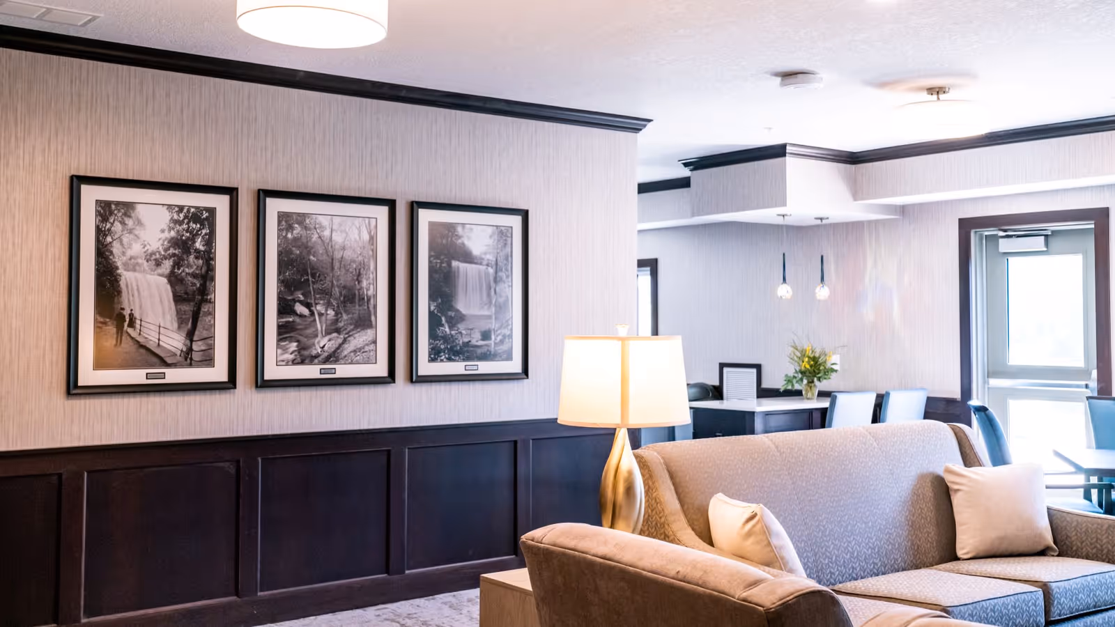 A cozy senior living common area with a beige couch adorned with light-colored pillows, a gold table lamp, and three framed black-and-white waterfall photographs on the wall. In the background, there is a dining area with tables and chairs near a door with a window.