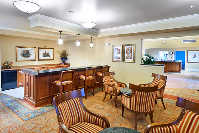 A cozy common area with a wooden bar counter and four bar stools. The room features several striped upholstered chairs arranged around small tables on a patterned carpet. The walls are decorated with framed artwork and there are pendant lights hanging above the bar. An open doorway leads to another room with additional seating and decor.