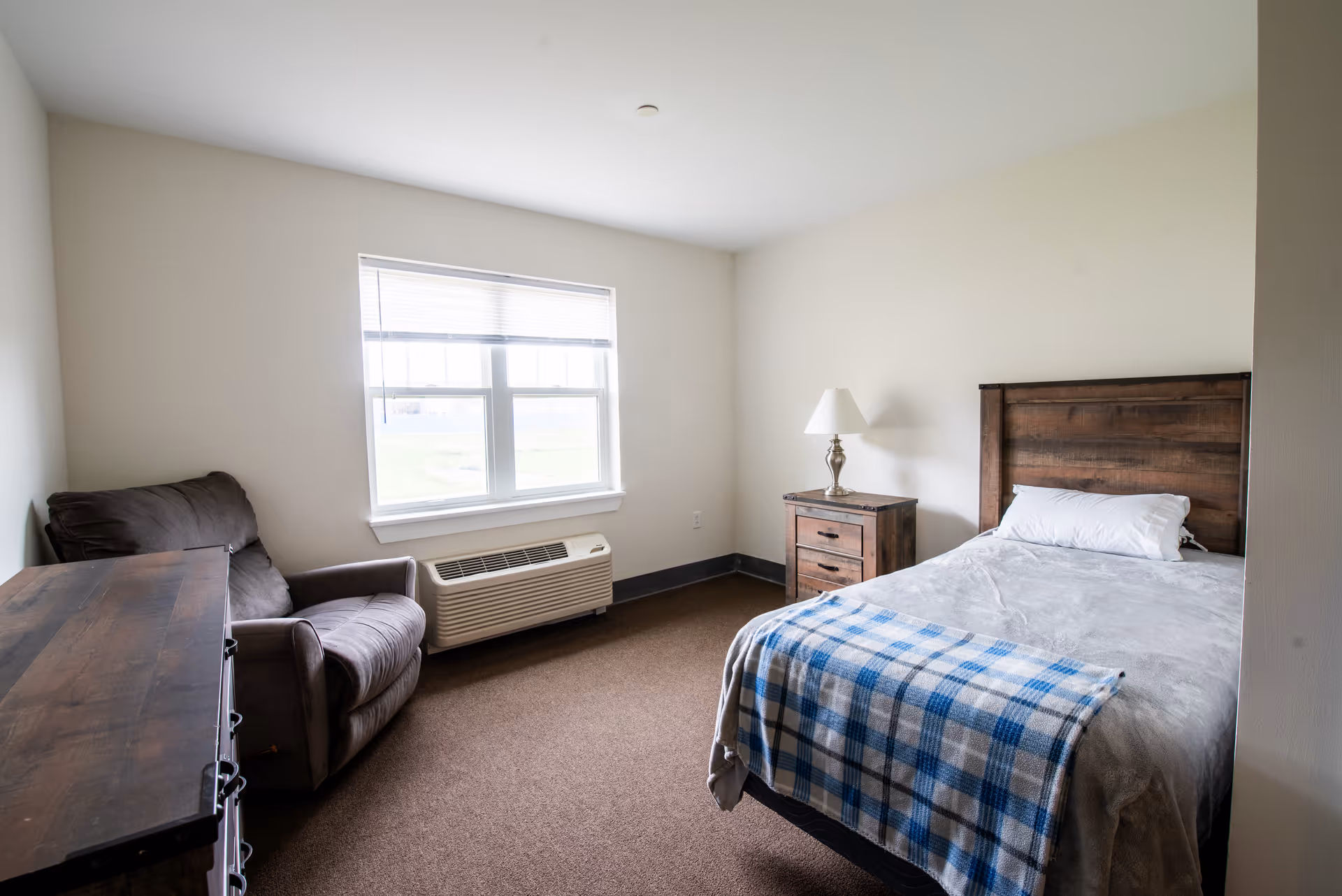 A simple bedroom with a wooden bed frame, a bed with a gray blanket and a blue plaid throw, a wooden nightstand with a lamp, a brown armchair, a wooden dresser, and a window with blinds letting in natural light.