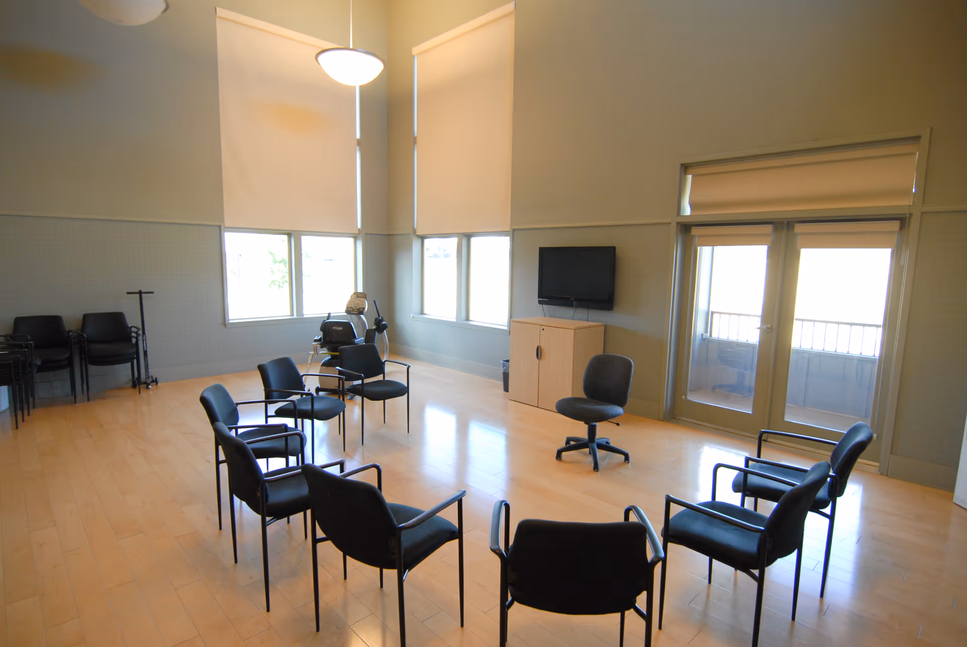 A spacious room with light wood flooring and high ceilings, featuring a circle of black chairs arranged in the center. There is a small cabinet with a wall-mounted TV above it, large windows with white blinds, and a glass door leading to an outdoor balcony. Additional stacked chairs and a coat rack are visible in the corner.