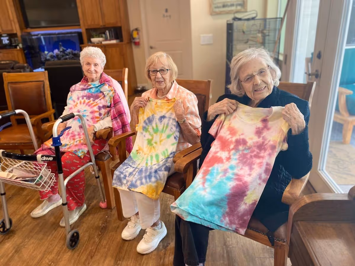 Three elderly women sitting in chairs inside a senior living facility, each holding up a colorful tie-dye shirt. One woman has a walker beside her, and the room has wooden flooring and furniture, with a glass door leading outside.