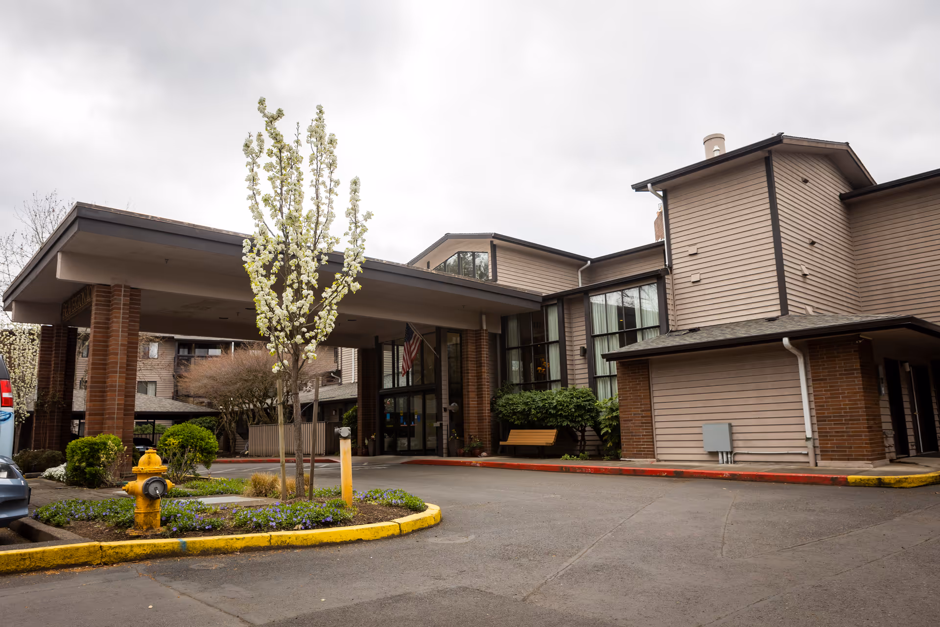 Exterior view of Beaverton Lodge senior living facility showing the entrance with a covered drop-off area, brick pillars, a small landscaped island with a flowering tree and a yellow fire hydrant, and a bench near the entrance under cloudy sky.