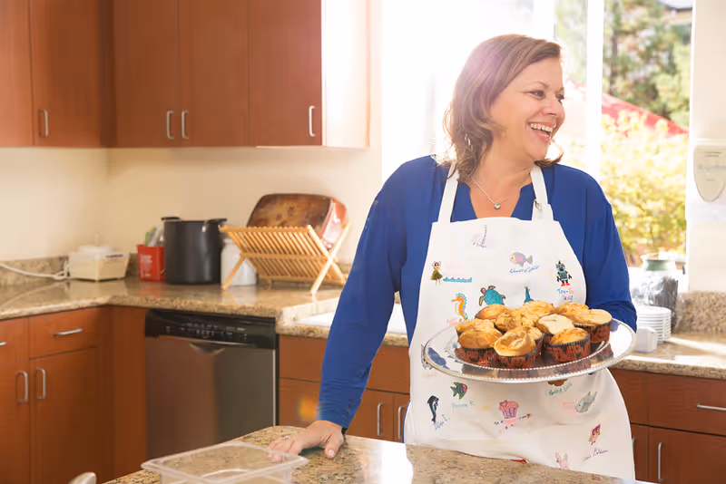 A woman wearing a white apron decorated with colorful drawings is standing in a kitchen holding a plate of muffins. She is smiling and looking to the side. The kitchen has wooden cabinets, a granite countertop, a dishwasher, and a dish rack near a window with sunlight coming through.