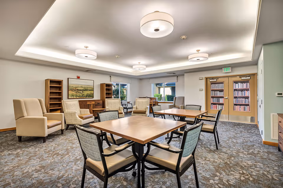 A well-lit common area with two square wooden tables surrounded by chairs in the foreground. In the background, there are several armchairs, bookshelves, a wooden cabinet, and a painting on the wall. Double doors with a bookshelf design are visible on the right side, and windows let in natural light.