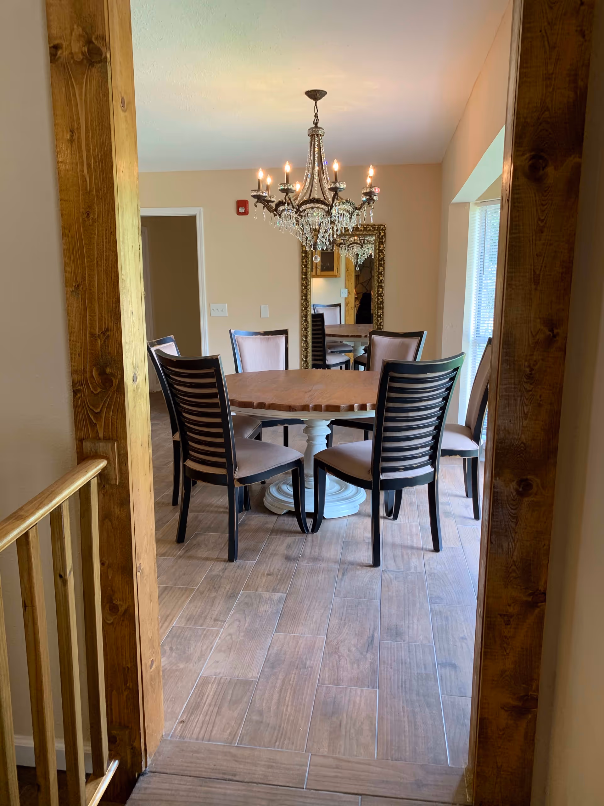 A dining room with a round wooden table surrounded by six chairs with cushioned seats. A decorative chandelier with lit candles hangs above the table. There is a large ornate mirror on the wall reflecting part of the room. The floor is covered with wood-like tiles, and the room is framed by wooden door trim.