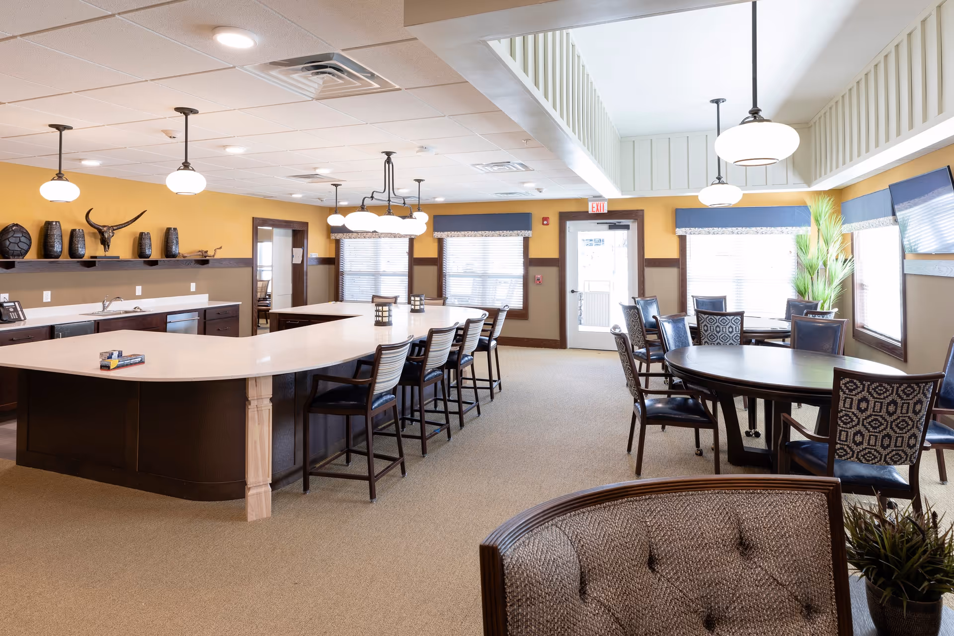 A bright and spacious dining area in a senior living facility featuring a large kitchen island with bar stools, round dining tables with patterned chairs, pendant lighting, and large windows with blinds. The walls are painted yellow with brown trim, and there are decorative vases and a mounted animal skull on a shelf. A TV is mounted on the wall near a potted plant.