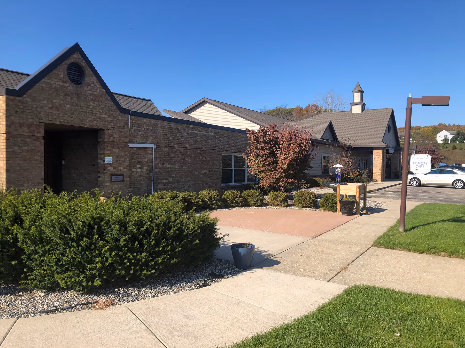 Front exterior of a single-story brick senior living building with landscaping, a walkway, parked cars, and a visible 'Memory Care' sign.