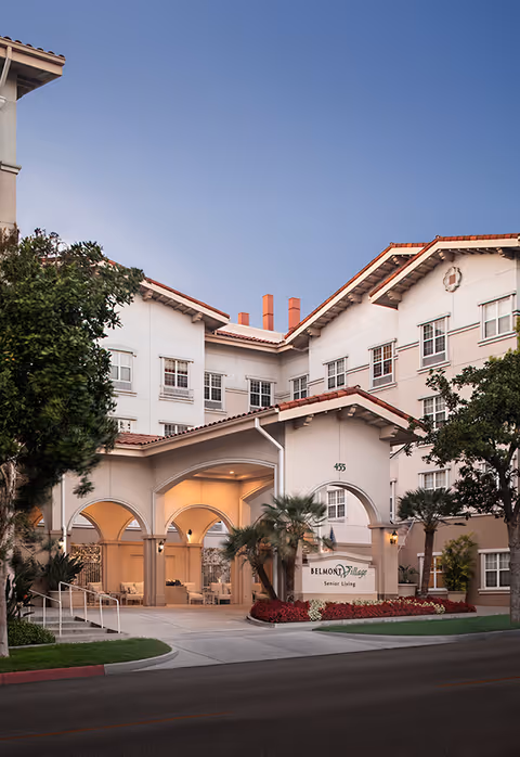 Exterior view of Belmont Village Senior Living Burbank building at dusk, featuring a covered entrance with arches, palm trees, and well-maintained landscaping.