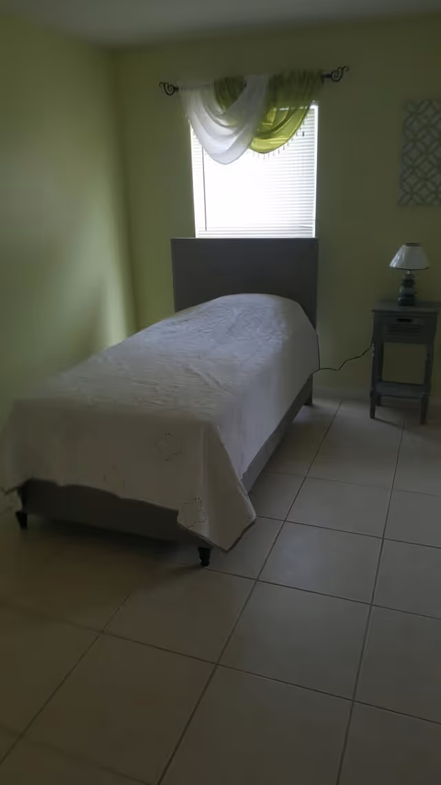 A small bedroom with a single bed covered with a white quilt. There is a window with white blinds and green and white sheer curtains above the bed. Next to the bed is a small gray nightstand with a lamp on it. The floor is tiled and the walls are painted light green.