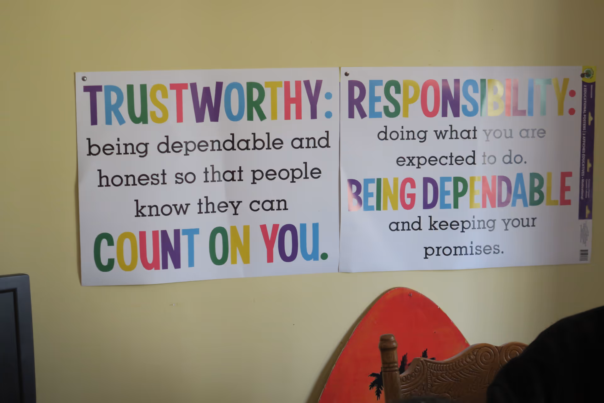 Two colorful posters on a yellow wall with motivational messages. The left poster reads: 'TRUSTWORTHY: being dependable and honest so that people know they can COUNT ON YOU.' The right poster reads: 'RESPONSIBILITY: doing what you are expected to do. BEING DEPENDABLE and keeping your promises.' A partial view of a wooden chair and a red surfboard-shaped object is visible below the posters.