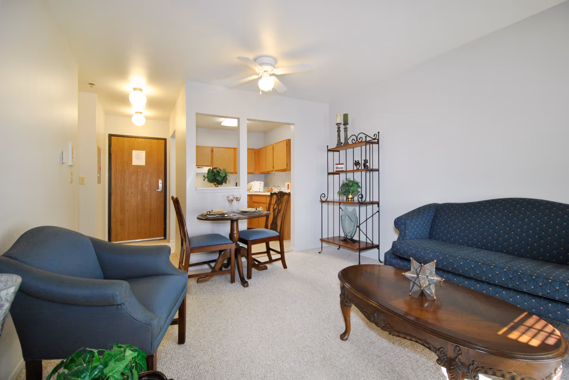 Well-lit living room with blue upholstered sofa and chair, wooden coffee table, small dining set, shelving unit, and an open view into the kitchen and entry door.