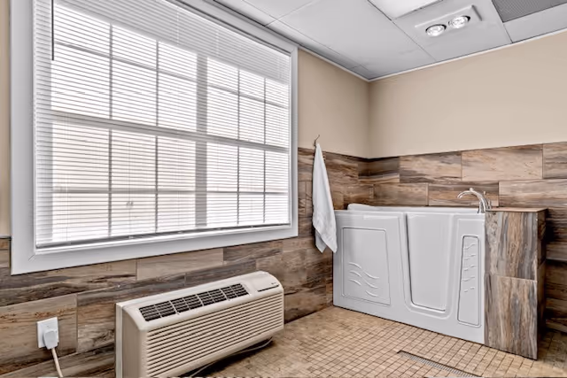 Small bathroom featuring a white walk-in tub, tiled walls and floor, a large window with blinds, a towel on a hook, and a floor heater unit.