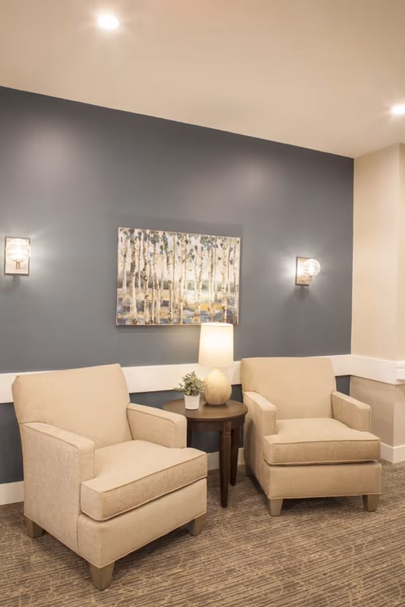 Two beige armchairs flanking a small round table with a lamp and artwork on a blue accent wall.