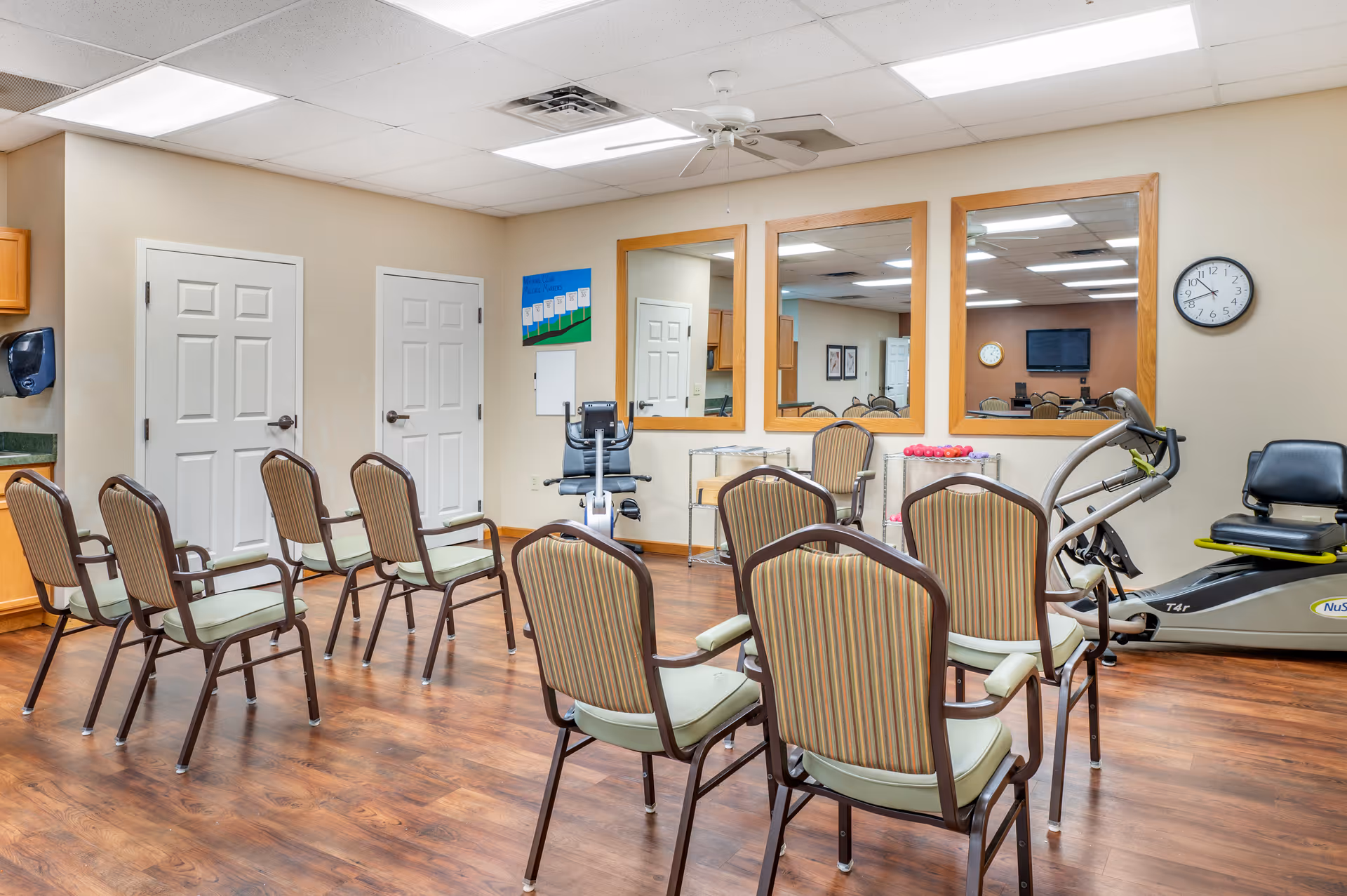 A room with wooden flooring and beige walls featuring two white doors, a row of chairs arranged in a semi-circle, exercise equipment including a recumbent bike and hand weights on a metal rack, three large mirrors on one wall, a wall clock, and fluorescent ceiling lights.