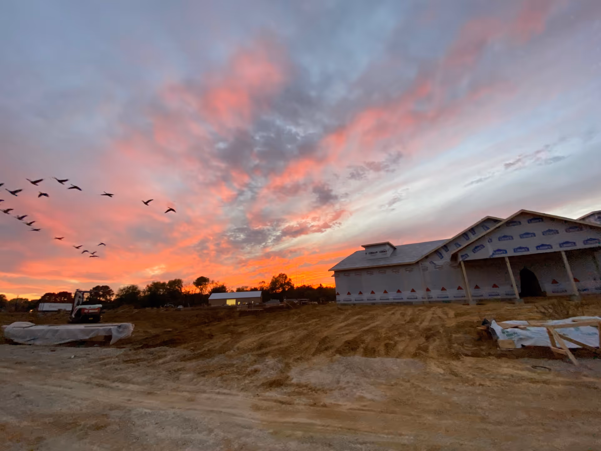A construction site at sunset with a partially built building covered in protective wrap. The sky is filled with vibrant orange, pink, and purple hues, and a flock of birds is flying across the sky. The ground is dirt with construction materials and equipment visible.