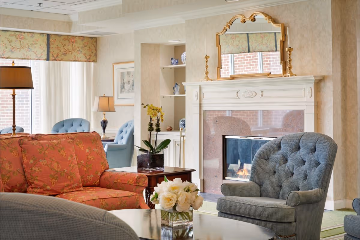 A bright, traditionally furnished living room with upholstered sofas and armchairs centered around a coffee table and a lit fireplace with a decorative mantel.